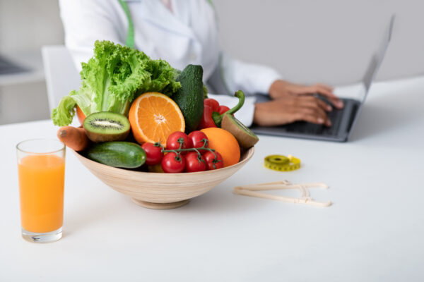 Cropped of black woman nutritionist doctor working with laptop at clinic, bowl with fruits and glass of juice on table, side view
